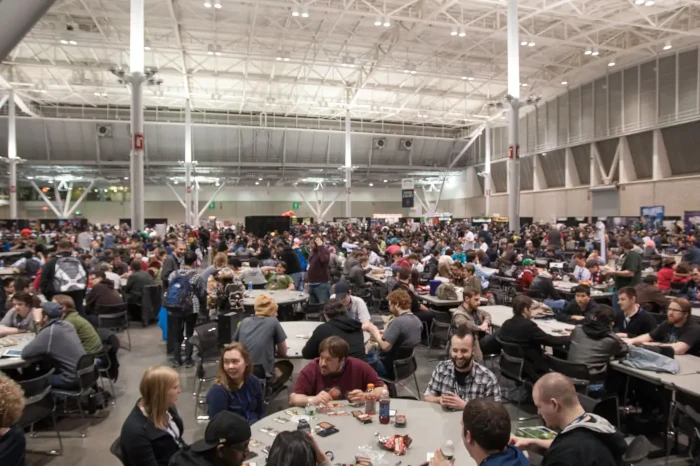 In a large white convention hall with a vaulted ceiling, many people are sitting or standing at a variety of gray tables, playing various games.