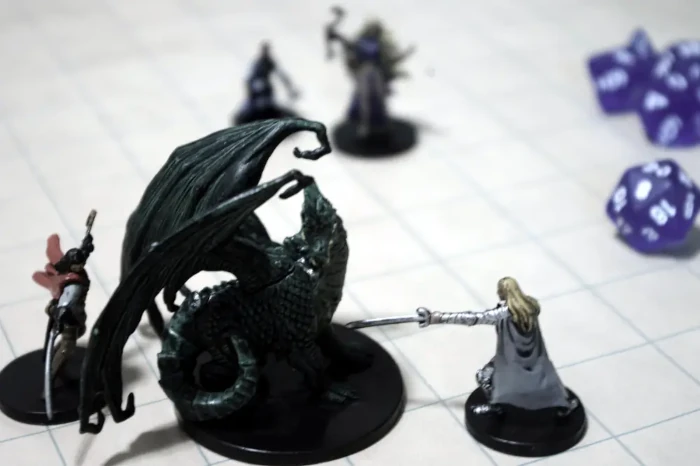 On a grid of squares are miniature figures and blue dice with white letters. In the foreground to the left, two male figurines wearing capes and armor are brandishing swords at a larger dark figure of a dragon set between them.