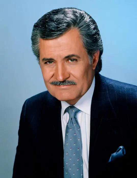 A mature man with well-styled gray hair and a trim mustache looks at the viewer with brown eyes and a slight smile. He sits in a dark blue suit against a light blue background.