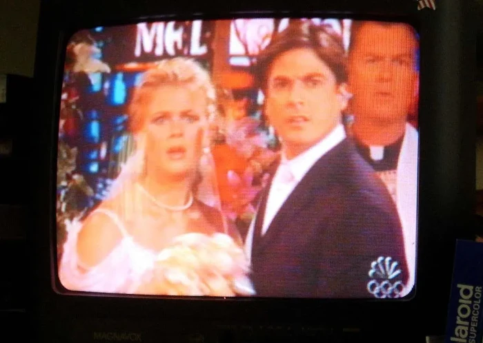 On a television screen is a wedding scene, but the blond bride and dark haired groom, as well as the priest behind them, are looking off-screen in shock.