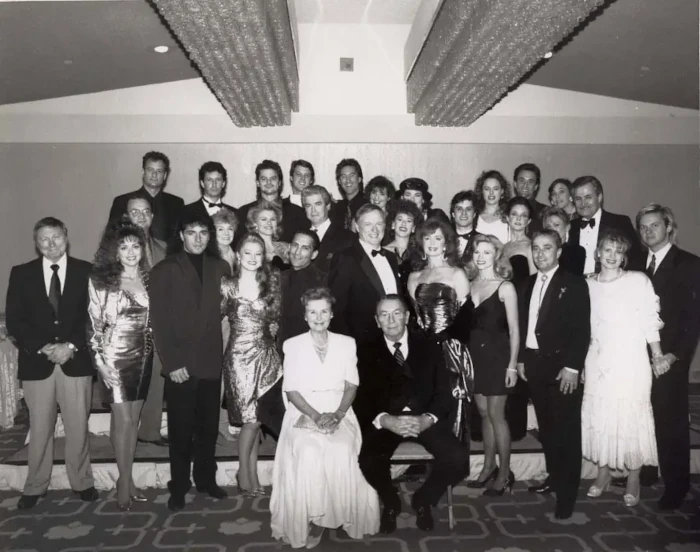 In this black and white photo, men in suits and women in dresses are arranged in three rows, with an older man and woman sitting on chairs at the front and center.